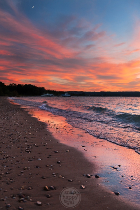 A vibrant sunset reflects off crashing waves at a pebble-strewn beach in Traverse City