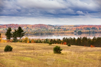 Warm fall colors in the hillsides around Lake Skegemog are doubled in reflection under moody skies